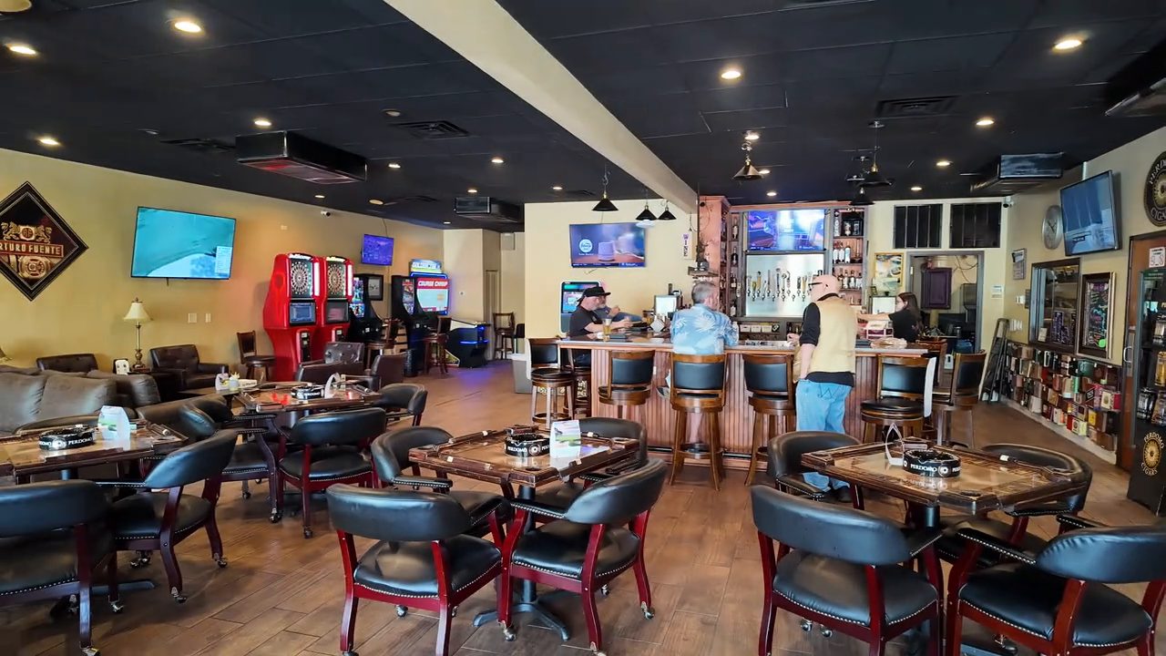 The main room of Island Girl Cigar Bar: round tables with black leather chairs in front, a long bar with stools at the back, and TVs on the walls.